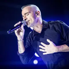 Wokalista, niczym prawdziwa **diva**, śpiewa z pasją, trzymając mikrofon i kładąc dłoń na sercu.