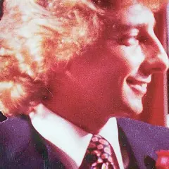 Uśmiechnięty mężczyzna w garniturze i krawacie, z burzą blond włosów. Atmosfera jak z **Studio 54**.