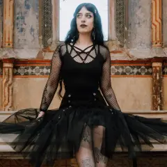 Kobieta w stylu trad goth siedzi na kamiennej ławie przed zniszczonym murem.
