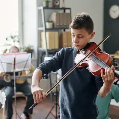 Chłopiec gra na skrzypcach z pasją, dźwięki rozbrzmiewają **fortissimo**. W tle inni uczniowie ćwiczą muzykę.