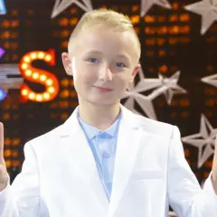 Chłopiec w białej marynarce i błękitnej koszuli pozdrawia, pokazując znak pokoju. Tło sugeruje scenę z Eurowizji Junior.