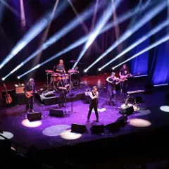 Zespół muzyczny na scenie, oświetlony niebieskimi i fioletowymi światłami. Wokalista na pierwszym planie, gitarzyści, skrzypek i perkusista. Prawdziwa uczta dla każdego melomana.