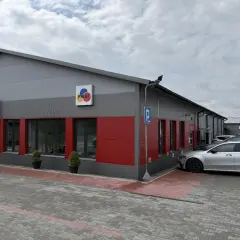 Nowoczesny budynek z czerwonymi akcentami i logo "PIRAT". Obok zaparkowane samochody, jeden srebrny, który wygląda jak z salonu, a nie jak bootleg.