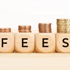 Cztery drewniane kostki z literami F, E, E, S tworzą napis FEES. Na każdej kostce stos monet, rosnący od lewej do prawej.