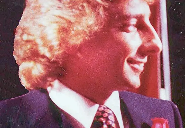 Uśmiechnięty mężczyzna w garniturze i krawacie, z burzą blond włosów. Atmosfera jak z **Studio 54**.