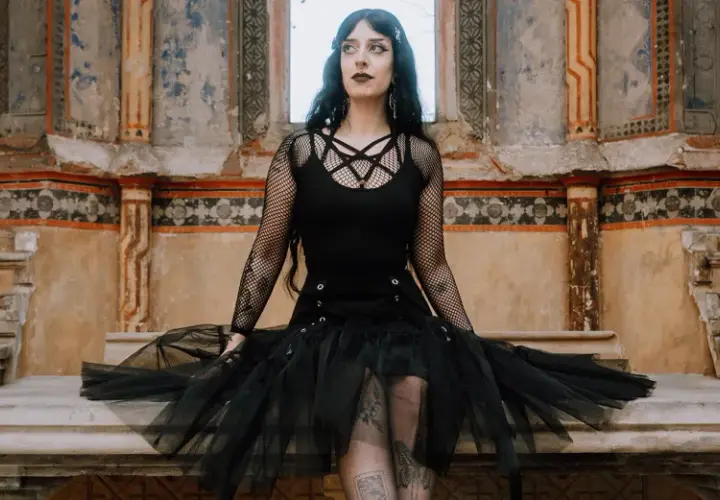 Kobieta w stylu trad goth siedzi na kamiennej ławie przed zniszczonym murem.