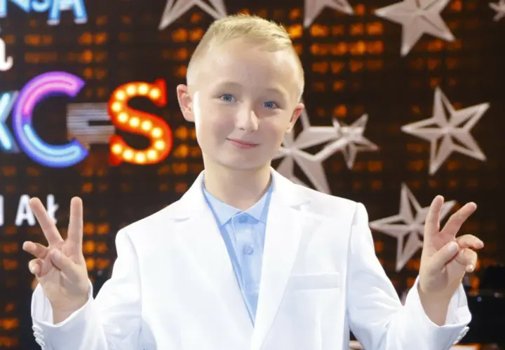Chłopiec w białej marynarce i błękitnej koszuli pozdrawia, pokazując znak pokoju. Tło sugeruje scenę z Eurowizji Junior.