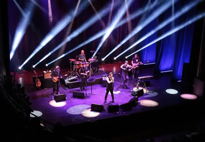 Zespół muzyczny na scenie, oświetlony niebieskimi i fioletowymi światłami. Wokalista na pierwszym planie, gitarzyści, skrzypek i perkusista. Prawdziwa uczta dla każdego melomana.