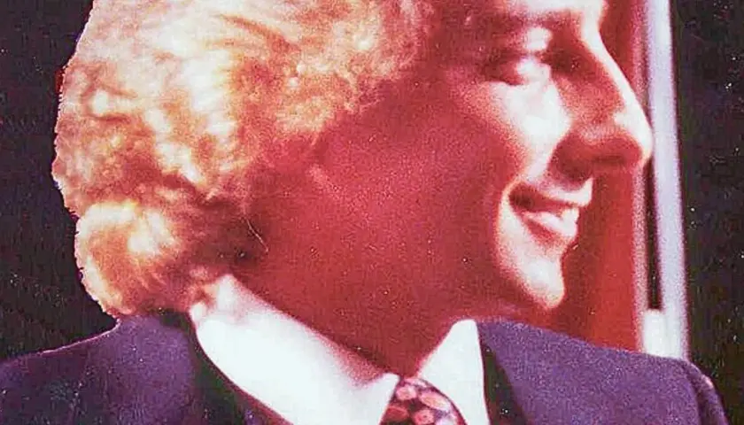Uśmiechnięty mężczyzna w garniturze i krawacie, z burzą blond włosów. Atmosfera jak z **Studio 54**.