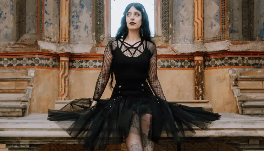 Kobieta w stylu trad goth siedzi na kamiennej ławie przed zniszczonym murem.