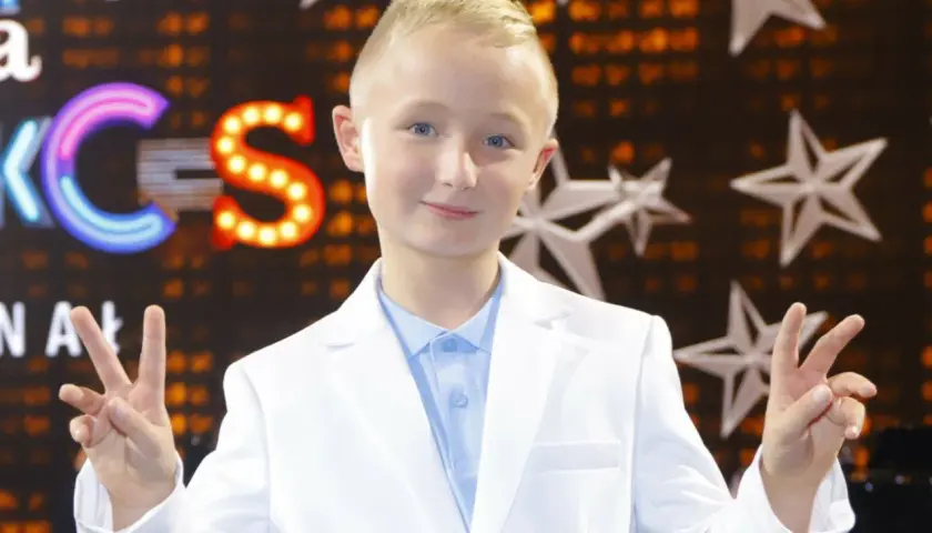Chłopiec w białej marynarce i błękitnej koszuli pozdrawia, pokazując znak pokoju. Tło sugeruje scenę z Eurowizji Junior.