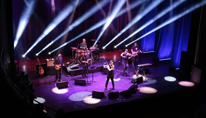 Zespół muzyczny na scenie, oświetlony niebieskimi i fioletowymi światłami. Wokalista na pierwszym planie, gitarzyści, skrzypek i perkusista. Prawdziwa uczta dla każdego melomana.