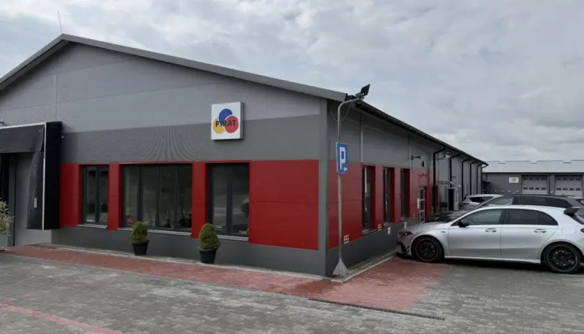 Nowoczesny budynek z czerwonymi akcentami i logo "PIRAT". Obok zaparkowane samochody, jeden srebrny, który wygląda jak z salonu, a nie jak bootleg.