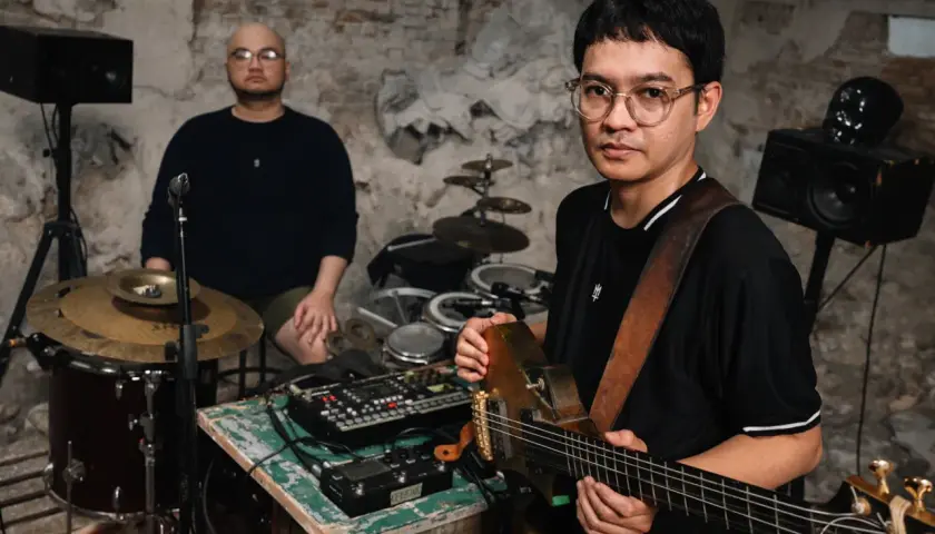 Dwóch muzyków w surowym wnętrzu, jeden gra na gitarze, drugi siedzi przy perkusji. Brzmienie tutti jest gotowe do nagrania.