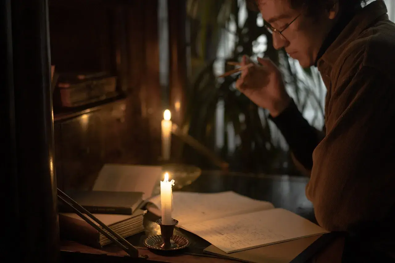 Mężczyzna w okularach pochyla się nad notatkami przy świecach, tworząc swoją własną, mroczną ballada.