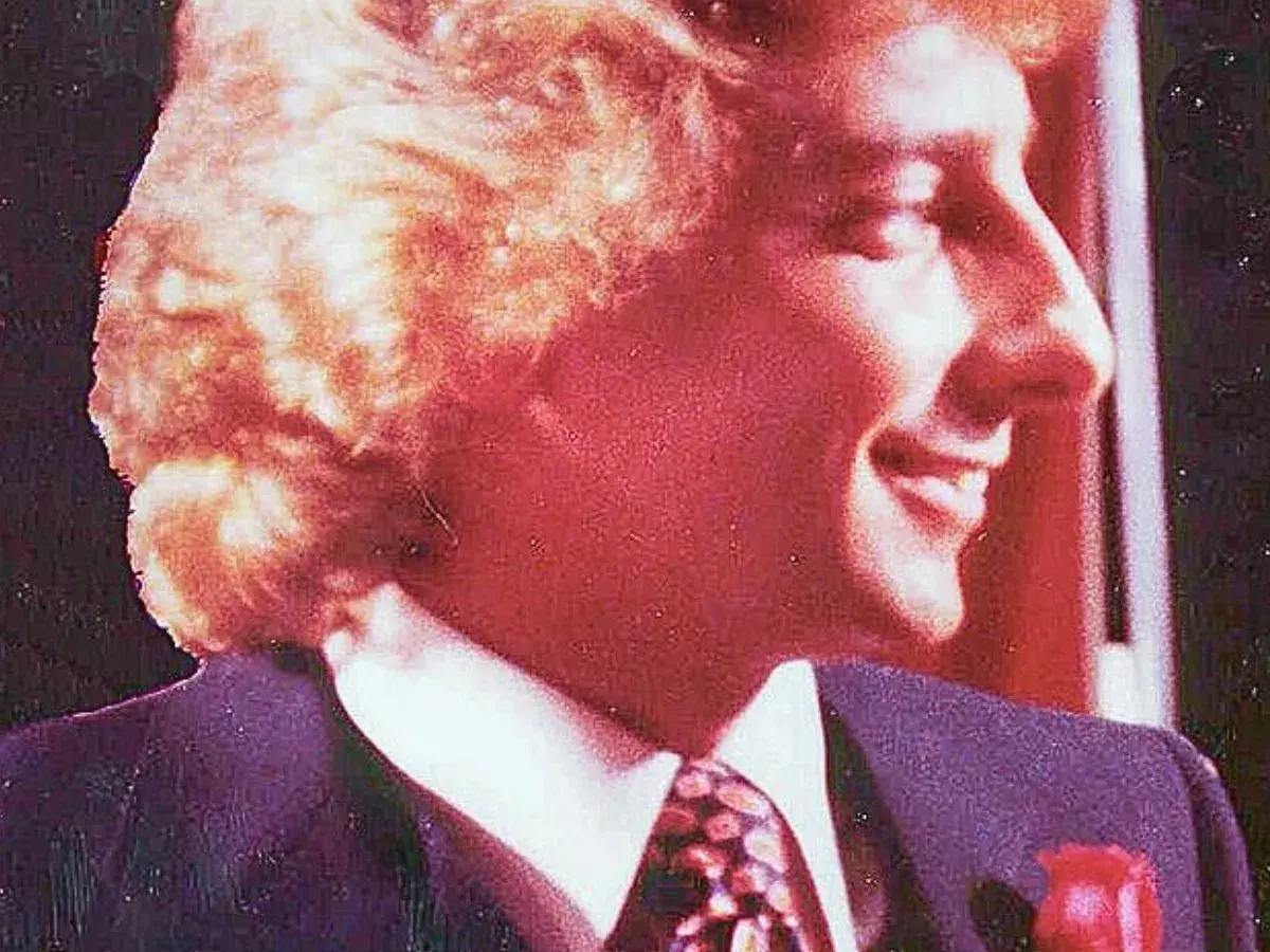 Uśmiechnięty mężczyzna w garniturze i krawacie, z burzą blond włosów. Atmosfera jak z **Studio 54**.