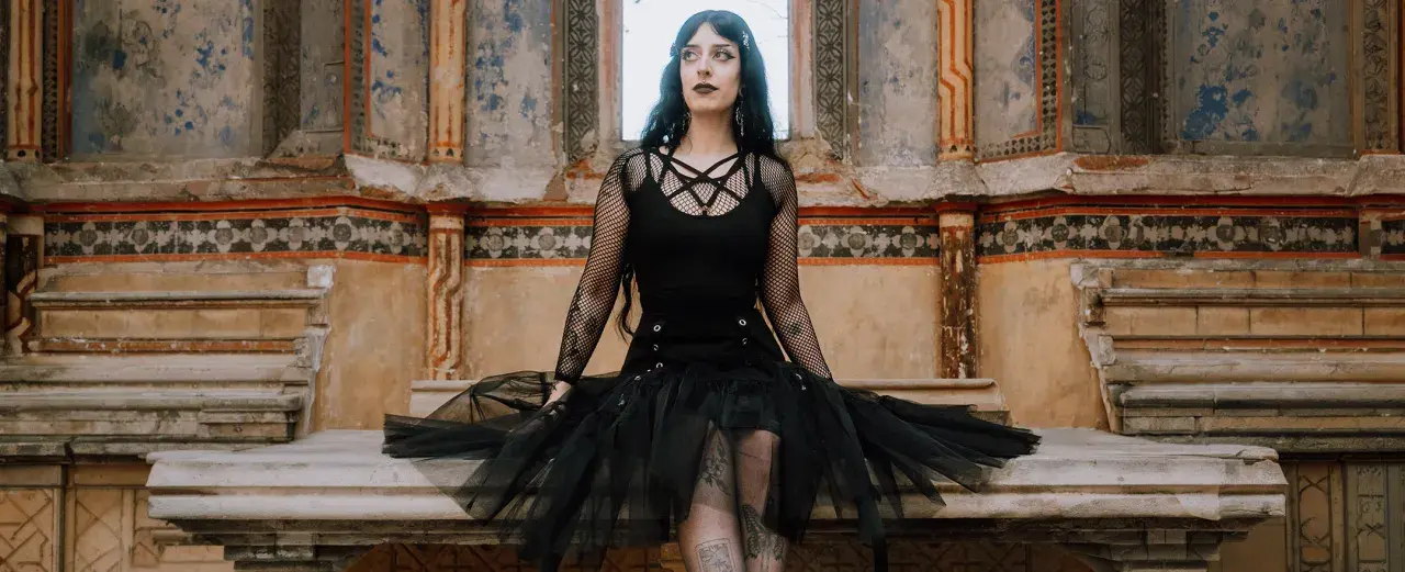 Kobieta w stylu trad goth siedzi na kamiennej ławie przed zniszczonym murem.