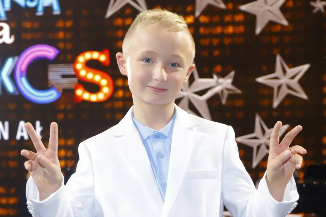 Chłopiec w białej marynarce i błękitnej koszuli pozdrawia, pokazując znak pokoju. Tło sugeruje scenę z Eurowizji Junior.