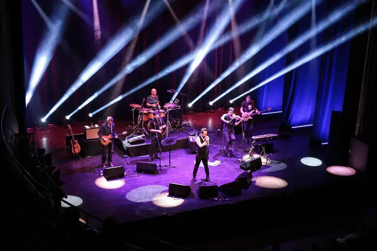 Zespół muzyczny na scenie, oświetlony niebieskimi i fioletowymi światłami. Wokalista na pierwszym planie, gitarzyści, skrzypek i perkusista. Prawdziwa uczta dla każdego melomana.