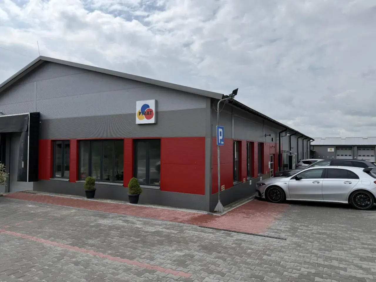 Nowoczesny budynek z czerwonymi akcentami i logo "PIRAT". Obok zaparkowane samochody, jeden srebrny, który wygląda jak z salonu, a nie jak bootleg.