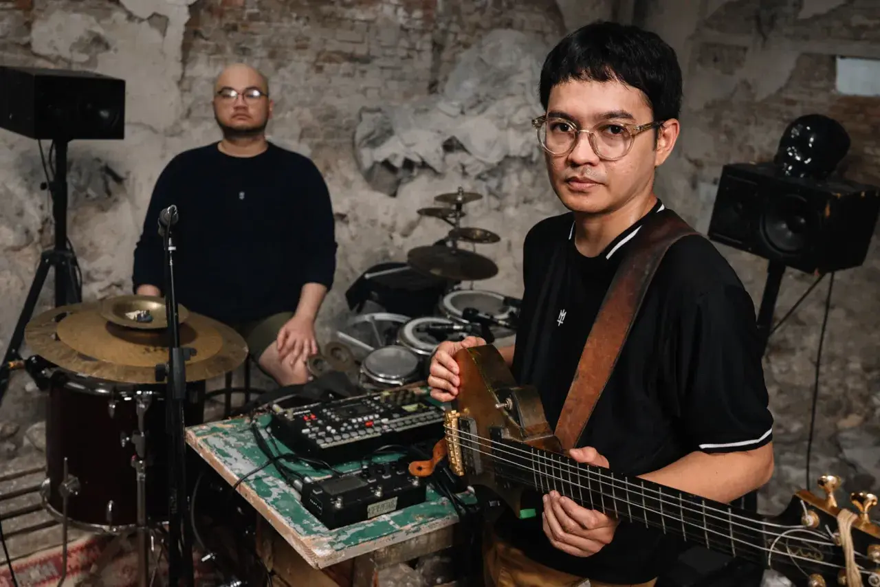 Dwóch muzyków w surowym wnętrzu, jeden gra na gitarze, drugi siedzi przy perkusji. Brzmienie tutti jest gotowe do nagrania.