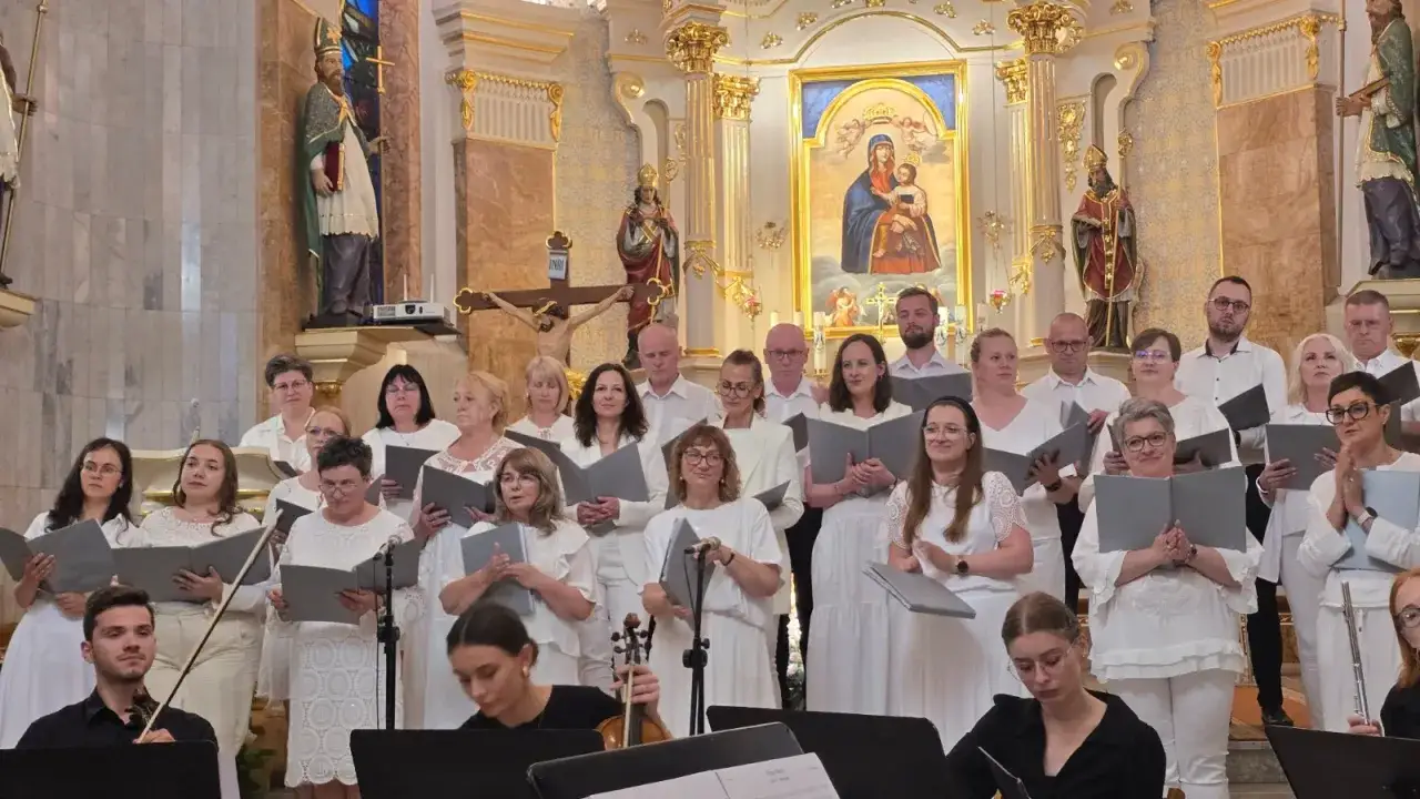 Chór w białych strojach śpiewa hymn w kościele, trzymając nuty. W tle ołtarz z obrazem Matki Boskiej i krucyfiksem.