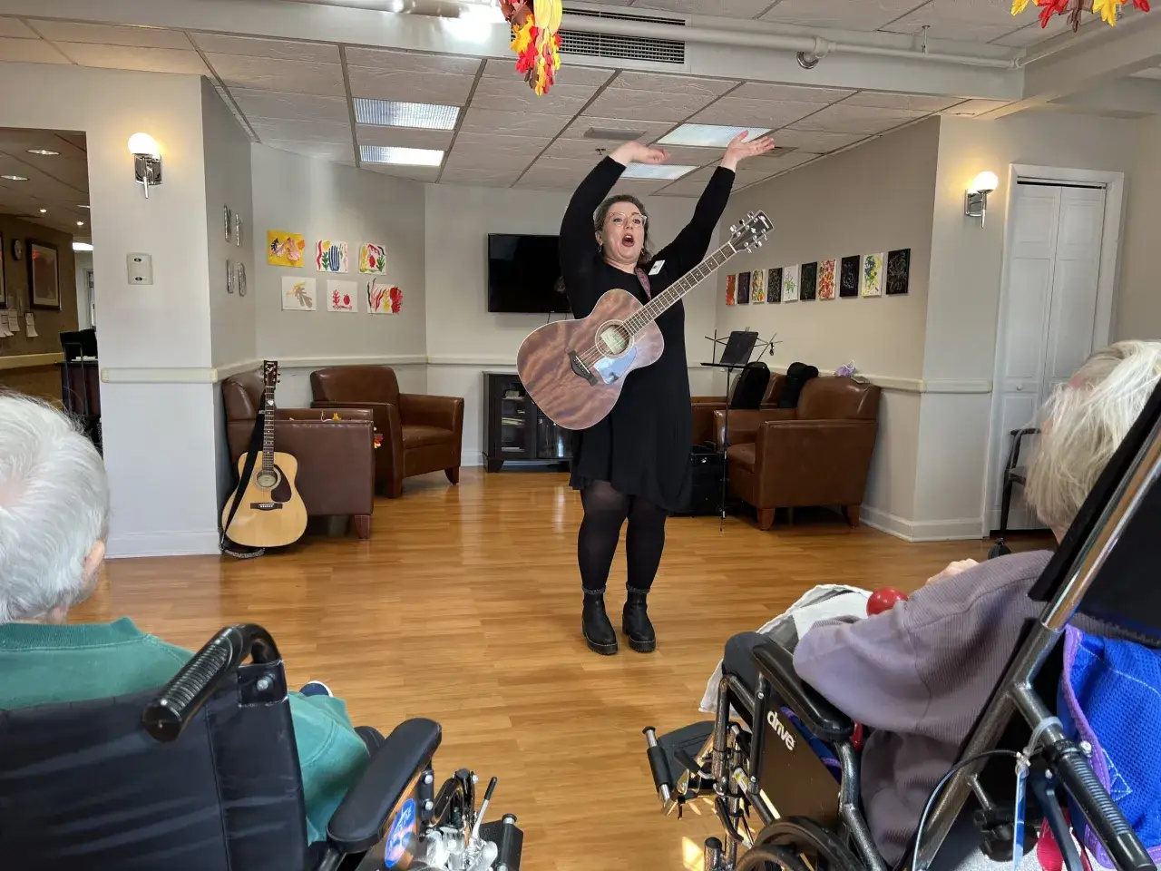 Kobieta z gitarą prowadzi sesję muzykoterapii dla seniorów na wózkach.